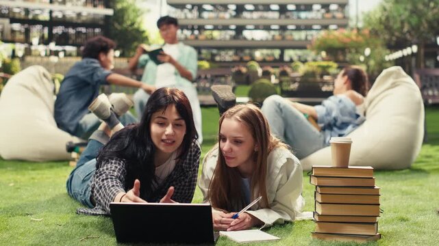 University students lie and lean over an open laptop and study materials on the grass, reviewing coursework in a textbook together during an outdoor peer tutoring session on campus.