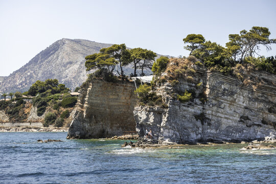 View of the rocky coastline with pine trees atop the cliffs and clear turquoise water in Zakinthos, Greece.