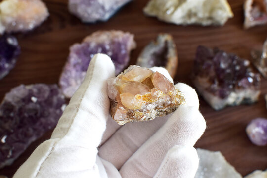A hand in a white glove holds an amethyst stone with crystals against a background of precious stones.