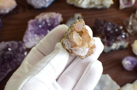 A hand in a white glove holds an amethyst stone with crystals against a background of precious stones.