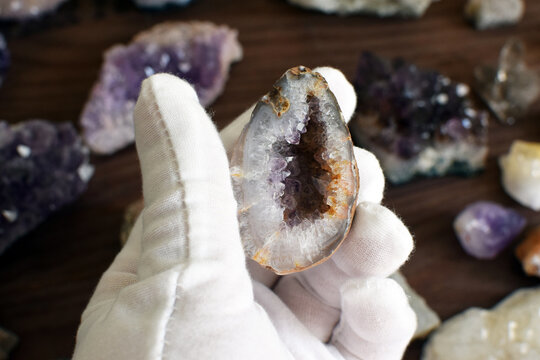A hand in a white glove holds an agate geode with amethyst crystals against a background of gemstones