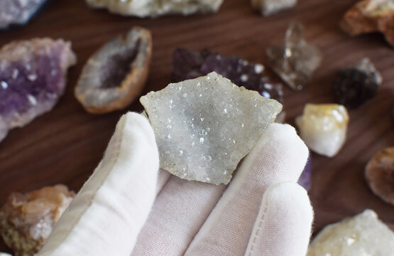 A hand in a white glove holds a rock with quartz made of small transparent crystals against a background of gemstones