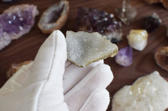 A hand in a white glove holds a rock with quartz made of small transparent crystals against a background of gemstones
