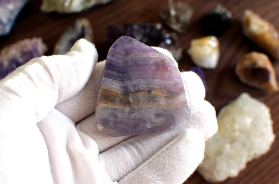 A white-gloved hand holds a polished striped purple fluorite against a background of gemstones