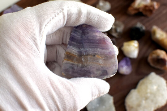 A white-gloved hand holds a polished striped purple fluorite against a background of gemstones