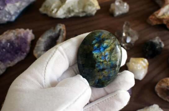 A hand in a white glove holds a tumbled labradorite stone with beautiful iridescence against a background of gemstones
