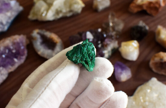 A hand in a white glove holds a green malachite stone against a background of gemstones