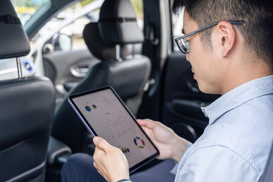 An adult Asian man wearing glasses sits in a car, holding a tablet and viewing data charts for work analysis.
