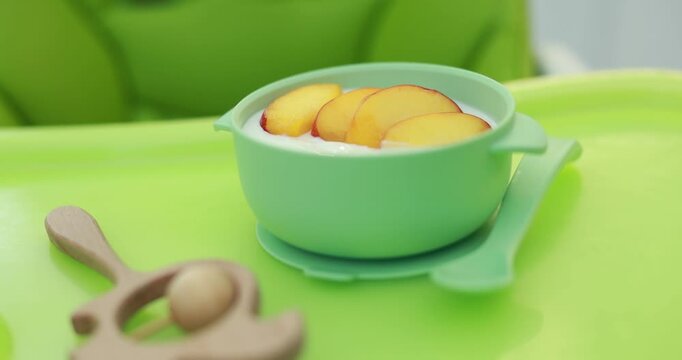 Baby tableware. Bowl with healthy food, spoon and teether on green feeding table, closeup. Camera moving forward
