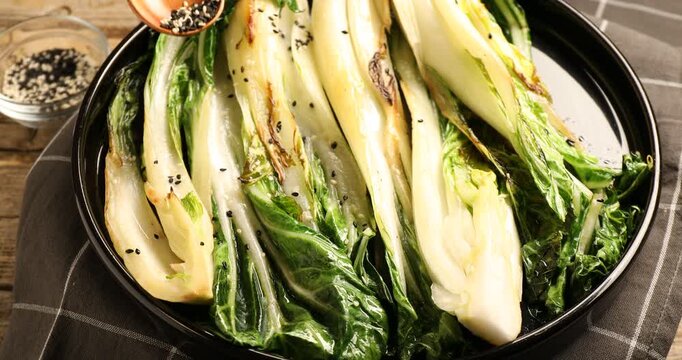 Putting sesame seeds onto tasty cooked bok choy cabbages on wooden table, closeup
