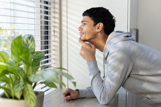African American male leaning on table near window blinds with plant, in gray hoodie, copy space