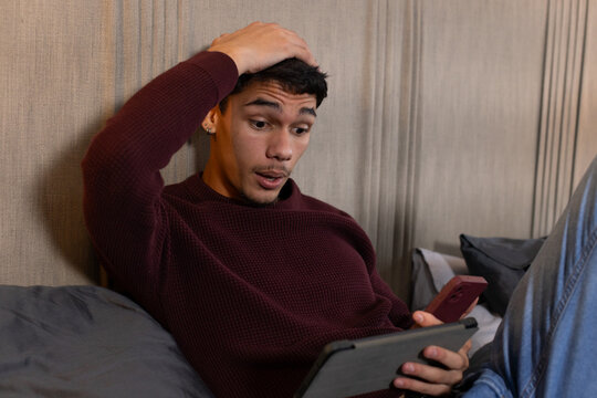 African American man reclining on bed, maroon sweater, holding tablet and red phone, raking hair