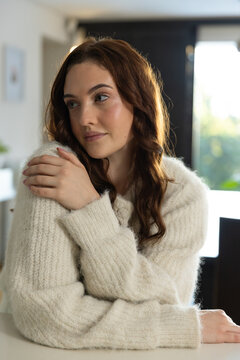 Woman sitting at table near large window wearing cream knit sweater with arms crossed