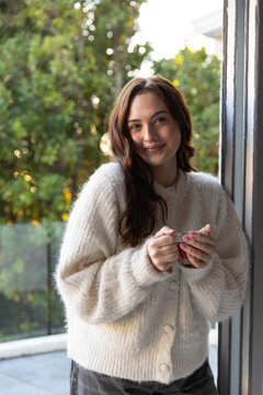 Smiling woman standing in doorway holding glass cup, wearing cream cardigan near glass railing