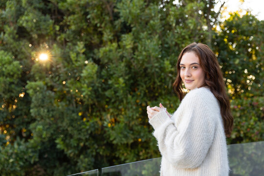 Adult woman standing on glass balcony holding small cup in fuzzy sweater, hedge sunbeam, copy space