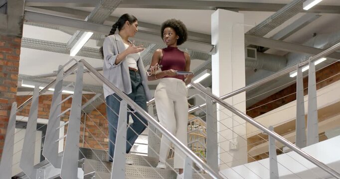 Diverse female coworkers stepping stairs at office, viewing tablet and phone for work as descending