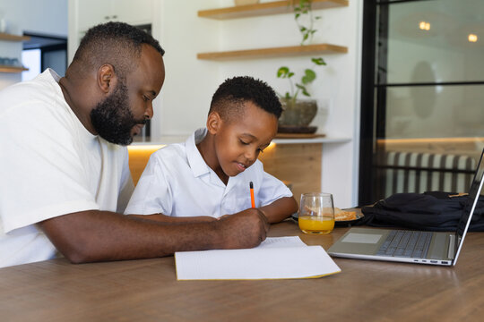 African American father and son tutoring at wood table in modern kitchen, writing on graph paper
