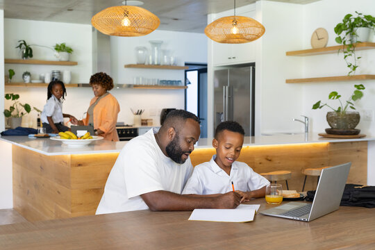 African American family studying in modern kitchen, father helping son with laptop and pencil