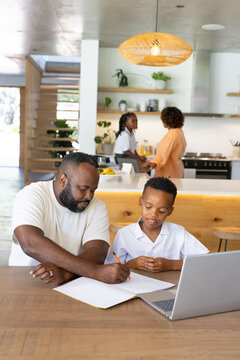 African American family working in open-plan kitchen, father guiding son with laptop and pencil