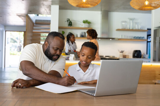 African family father helping youth son in white shirt with notebook at open-plan kitchen laptop