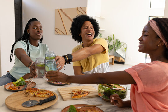 African American women sitting at home table sharing pizza, salad, clinking cucumber water glasses