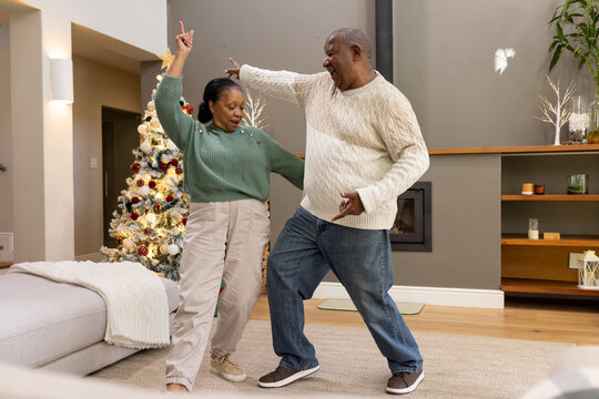 Senior woman dancing with African American man in living room wearing sweaters by Christmas tree