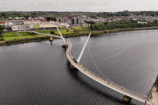 Aerial view of the Peace Bridge spanning the River Foyle with pedestrians, surrounded by urban buildings and green landscapes in DerryLondonderry, Northern Ireland, United Kingdom.