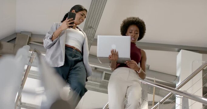 Diverse women coworkers walking down stairwell reviewing brief on laptop and phone in office attire