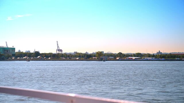 Coastline of Governors Island viewed from a moving boat. A distant shore with green trees and white tents sits across calm water under a bright sky.