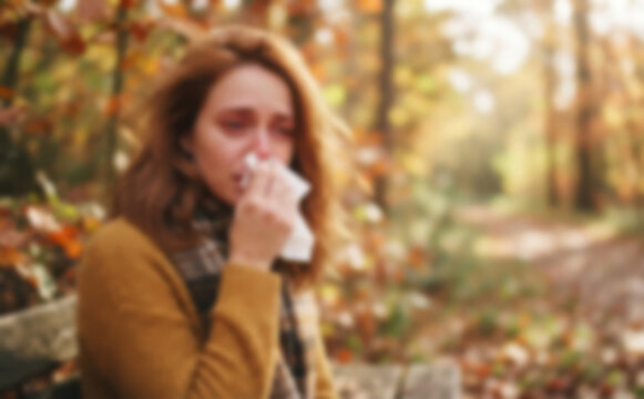 Young adult woman blowing her nose with a tissue in an autumn park, concept of seasonal allergies and cold illness