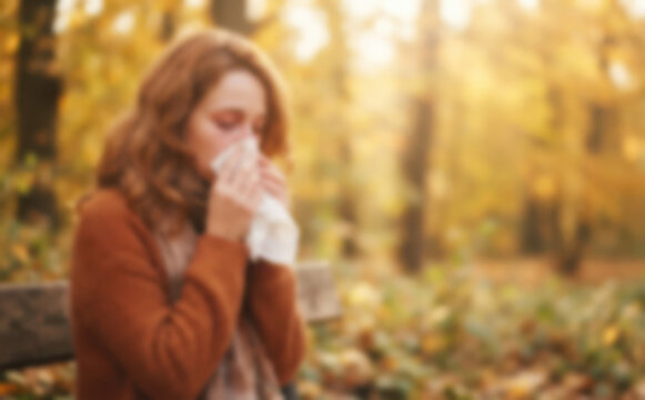 Young adult woman sneezing into a tissue in an autumn park, suffering from seasonal allergies or cold outdoors, health issue concept