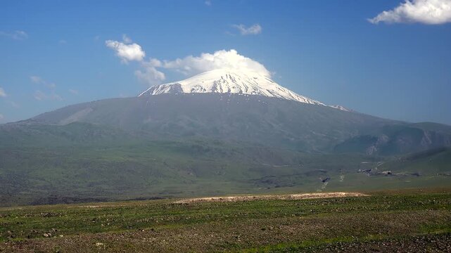 Mount Ararat snowy summit in Turkey rises above green plain as clouds drift in time lapse sequence.