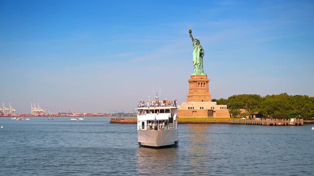 New York, USA, 4 Jule 2026 : Tourist ferry moving on the river near the Statue of Liberty. A white deck boat carrying passengers sails toward the iconic monument under a blue sky.