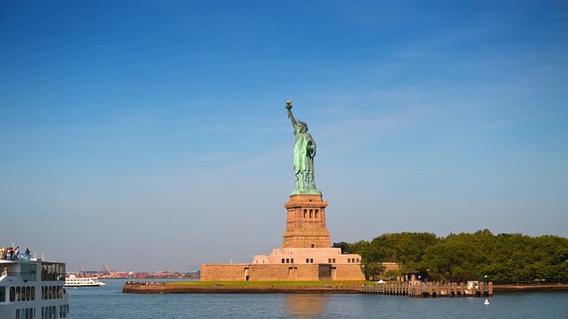 Statue of Liberty standing under a clear blue sky in New York. The green copper monument rises from its stone pedestal near green trees on a sunny day.