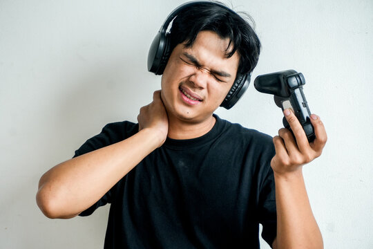 Asian Man Wincing in Pain While Massaging Neck Holding Game Controller Wearing Headphones