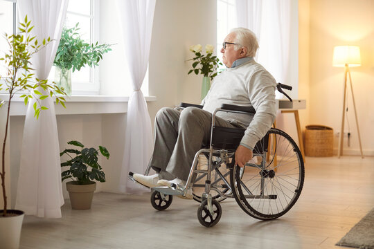 Senior elderly man sitting in wheelchair, looking pensively through window, suffers from health problems, has physical, feels lonely at home. Old people and retirement 
