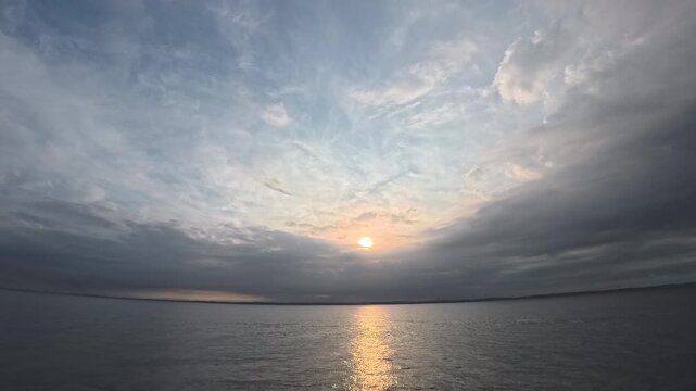 Cinematic real-time wide angle video of gentle sun breaking through layered clouds over calm ocean with golden reflection path