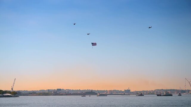 Helicopters carrying an American flag over the harbor. Military aircraft fly in formation above the water during a warm sunset celebration in New York.