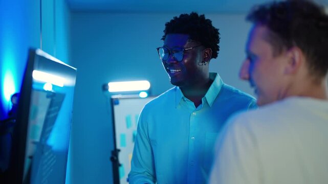 Two diverse IT professionals collaborate in a dark server room. Smiling male software engineers discuss data on a computer monitor, pointing at code in a blue neon lit modern technology office.