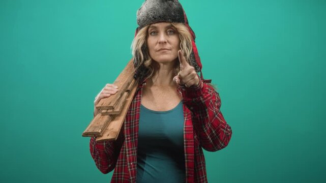 Woman in fur trapper hat and plaid shirt carrying wooden planks on shoulder points finger at camera in studio; determination warning resilience.