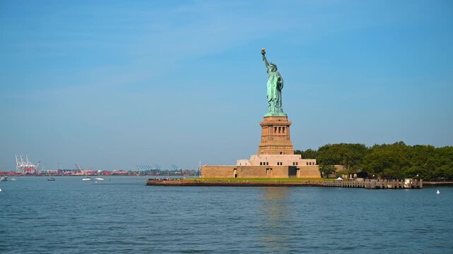Statue of Liberty standing on Liberty Island. The famous copper monument rises against a clear blue sky surrounded by green trees and calm water.
