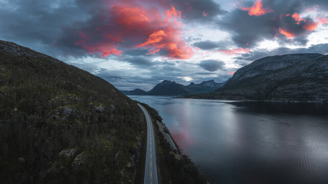 Aerial view of the iconic Stetind mountain rising above a winding road along the fjord under a dramatic sunset sky in Narvik Municipality, Nordland, Norway.