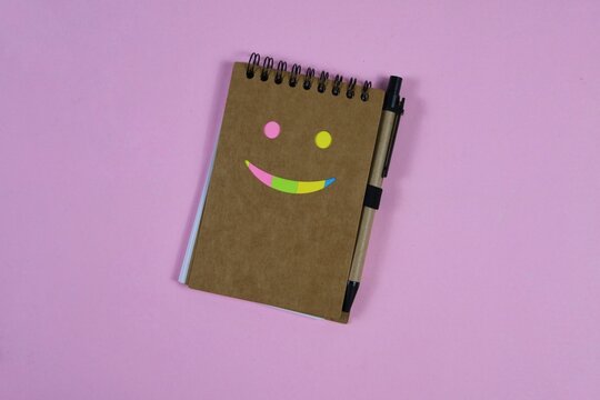 Colorful smiley face made with sticky notes on a spiral notebook and pen, arranged on an pink background with ample copy space.