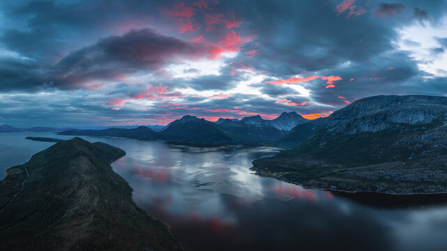 Aerial view of the majestic Stetind mountain rising above the calm waters of the fjord under a dramatic sunset sky with vibrant pink and blue clouds in Narvik Municipality, Nordland, Norway.