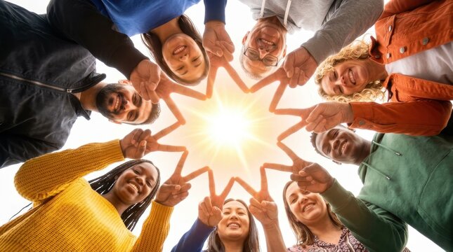 Happy group of diverse multicultural people looking down at camera forming a star shape with fingers outdoors against bright sun, showcasing friendship, unity, community, and global peace