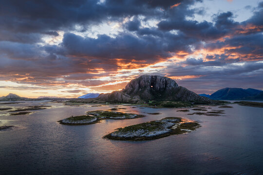 Aerial view of Torghatten mountain with its iconic natural tunnel, surrounded by coastal waters and small islands under a dramatic sunset sky in Bronnoy Municipality, Nordland, Norway.