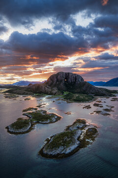 Aerial view of Torghatten mountain with its iconic natural tunnel, surrounded by rocky islands and calm coastal waters under a dramatic sunset sky in Bronnoy Municipality, Nordland, Norway.