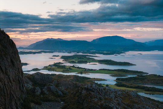 Aerial view of Torghatten mountain and surrounding coastal islands with lush green fields and calm waters under a dramatic sunset sky in Bronnoy Municipality, Nordland, Norway.