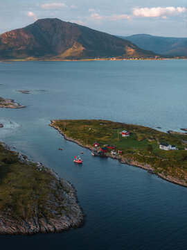 Aerial view of Torghatten mountain rising above a small island with traditional red cabins and a fishing boat in the coastal waters of Bronnoy Municipality, Nordland, Norway.