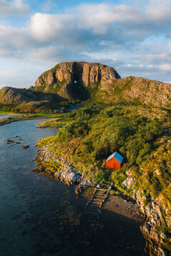 Aerial view of Torghatten mountain with its iconic natural tunnel, featuring a red wooden cabin by the rocky coastline and lush green landscape in Bronnoy Municipality, Nordland, Norway.